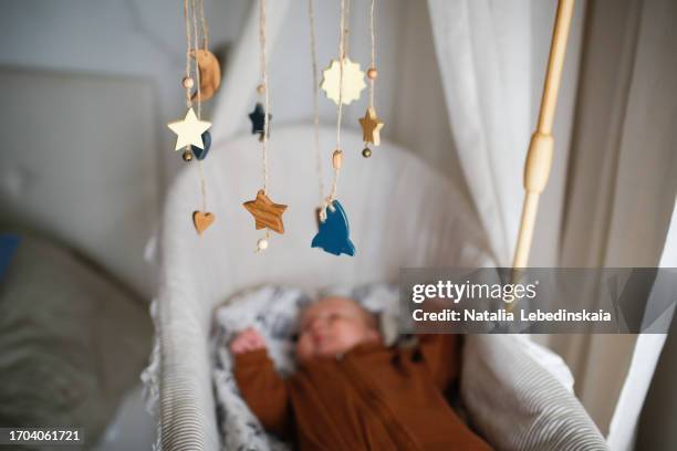 eco-conscious infancy: baby, dressed in brown overalls, lies in a bassinet, gazing at a wooden baby mobile, symbolizing mindful and sustainable toy selection. - baby mobile stock pictures, royalty-free photos & images