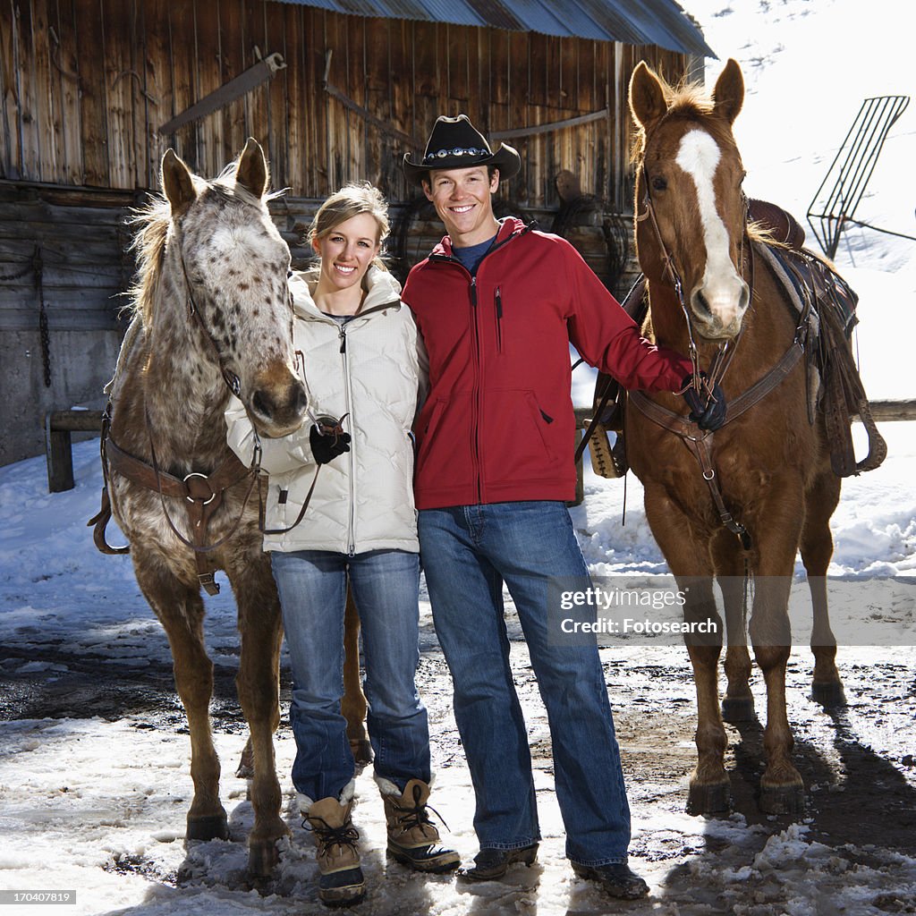 Couple holding horses in winter with stable in background