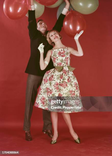 Posed studio portrait of a female fashion model wearing a sleeveless poppy print dress with bow detail and full skirt, she stands with a man as they...