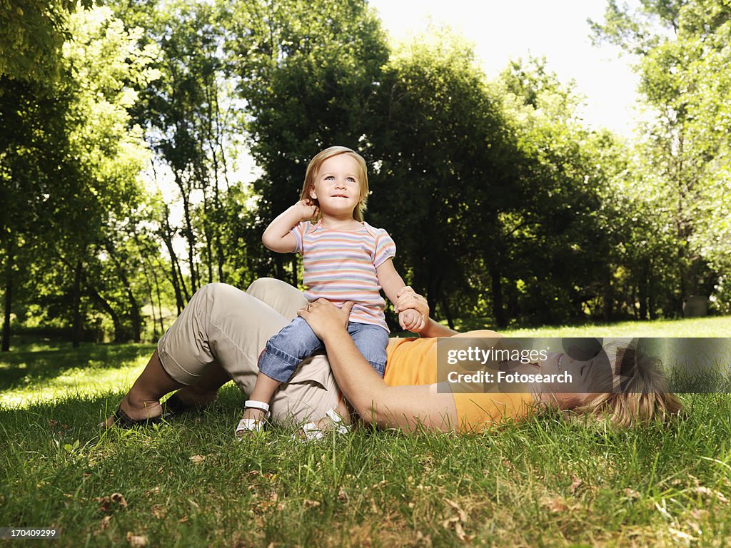 Woman lying in grass at park with toddler daughter seated on her lap
