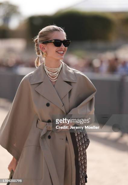 Xenia Adonts is seen outside Dior show wearing black Dior sunnies, golden hoop earrings, layered Dior necklace, beige Dior trench coat, beige colored...