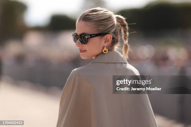 Xenia Adonts is seen outside Dior show wearing black Dior sunnies, golden hoop earrings, layered Dior necklace, beige Dior trench coat, beige colored...