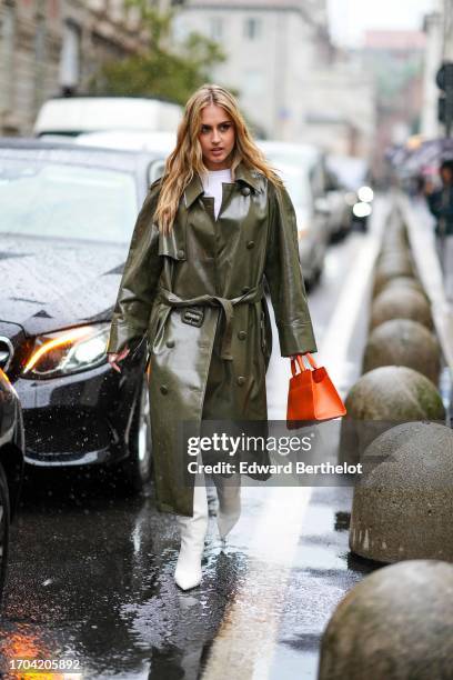Guest wears a khaki green double-breasted raincoat, white pointy high-heeled boots, an orange leather bag, outside Aigner, during the Milan Fashion...