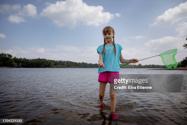 girl (10-11 years) wading through the shallow waters near the shore of a lake on a beautiful summer's day, holding a toy fishing net whilst offering forwards a found shell in her hand. - 10 11 years stock pictures, royalty-free photos & images