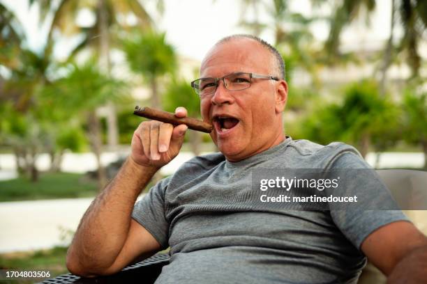 handsome 50+ man smoking a cigar in a tropical setting. - cigar stock pictures, royalty-free photos & images