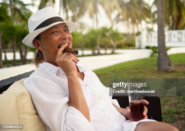 beautiful 50+ woman smoking a cigar in a tropical setting. - cigar stock pictures, royalty-free photos & images