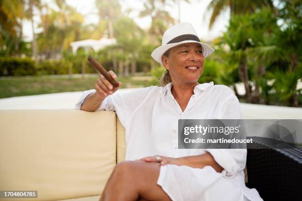 hermosa mujer 50+ fumando un cigarro en un entorno tropical. - beautiful women smoking cigars fotografías e imágenes de stock