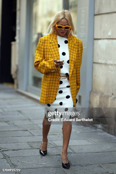 Tina Leung wears yellow sunglasses, a checked oversized blazer jacket, a white and black polka dots printed on-knee dress, pointed black shoes,...