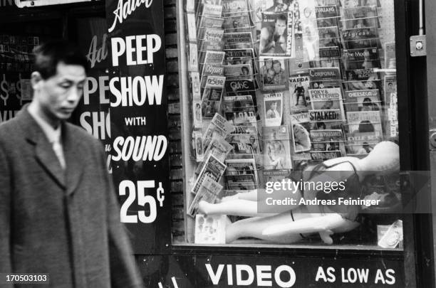 Man walking past the window of a sex shop in New York City, circa 1980. The window display features a range of pornographic magazines, along with a...