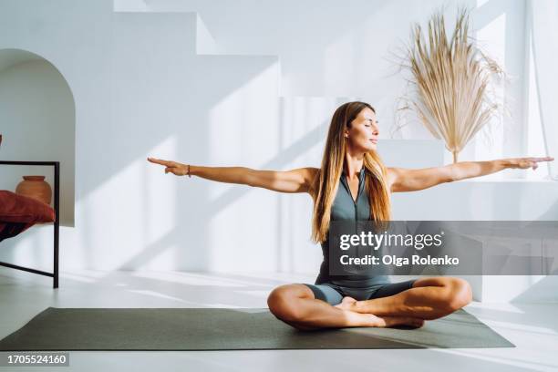 relaxed blonde woman with arms outstretched, smile to morning sunlight during breathing exercises on mat at home - cross legged stock pictures, royalty-free photos & images