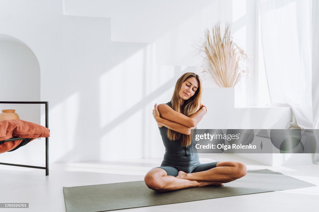 Yoga self care in the morning: blonde woman embracing herself, practicing breath work in white room in the morning