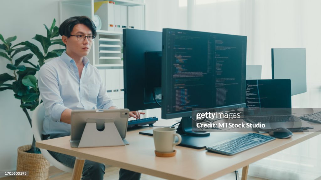 Young Asian man software developers using computer to write code sitting at desk with multiple screens work at office. Programmer development.