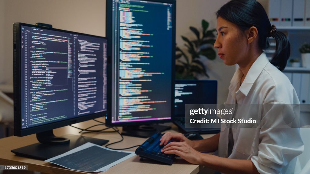 Young Asian woman software developers using computer to write code sitting at desk with multiple screens work in office at night. Programmer development.