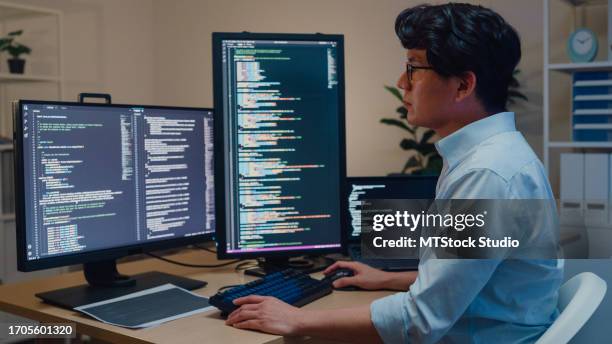 young asian man software developers using computer to write code sitting at desk with multiple screens work in office at night. programmer development. - application programming interface stockfoto's en -beelden