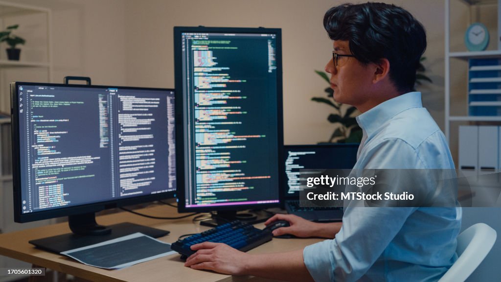 Young Asian man software developers using computer to write code sitting at desk with multiple screens work in office at night. Programmer development.