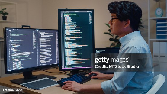 Young Asian man software developers using computer to write code sitting at desk with multiple screens work in office at night. Programmer development. Young Asian man software developers using computer to write code sitting at desk with multiple screens work in office at night. Programmer development.
