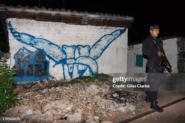 Heavily armed guard keeps watch for gang related activity in front of an exterior graffitied with the Devils Head, an MS-13 insignia in the...