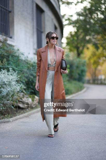 Darja Barannik wears sunglasses, a brown long trench leather coat, a crop top / low-neck top, gray flared pants, outside Christopher Esber, during...