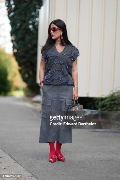 Guest wears red suglasses, long earrings, a v-neck sleeveless gray wool top with blue details, a midi skirt with pockets, a gray Hermes bag, red...