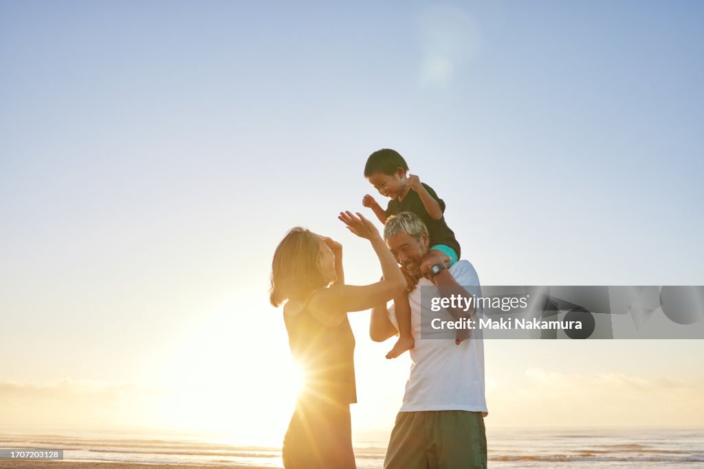 Father carrying an infant on his shoulders on the shore at sunrise, and a mother beside him.