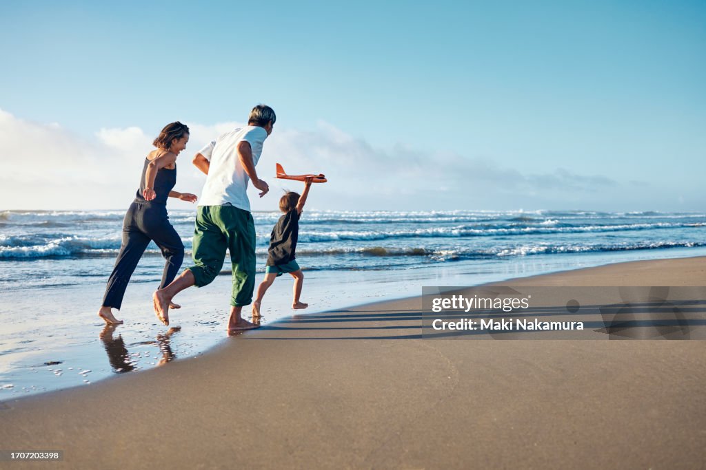 Parents running behind a boy running straight ahead with a model airplane held high in one hand.