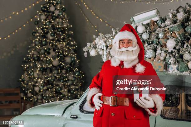 happy santa claus holding a christmas gift in his hand against the background of a christmas car looking at the camera - machine christmas tree stock pictures, royalty-free photos & images