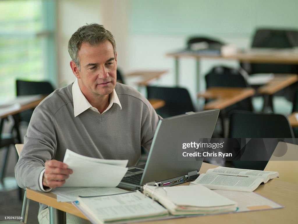 Professor using  laptop with reveiwing paperwork in classroom