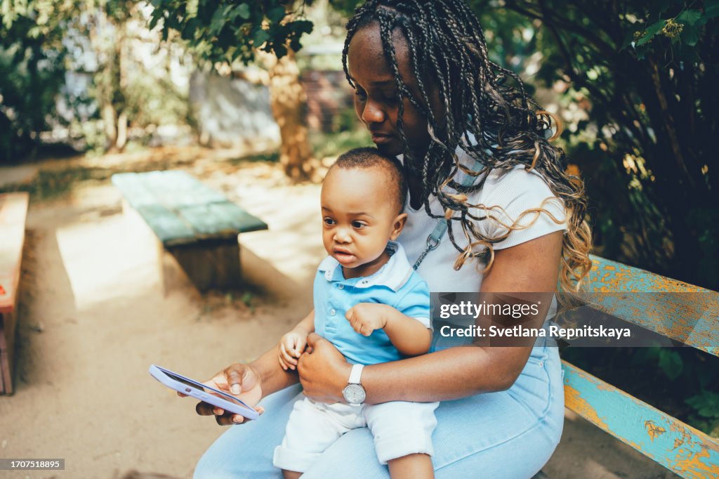 African mother using smartphone with child while sitting on a bench on the street in summer