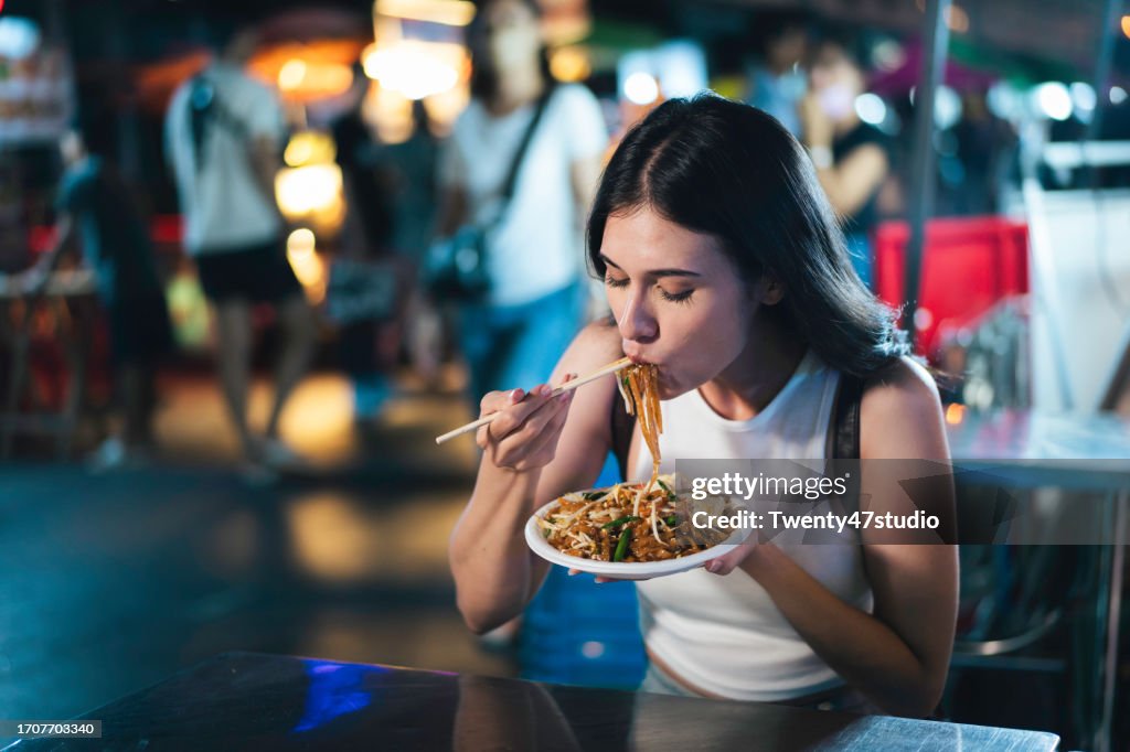 A young woman eating Pad Thai while traveling on Khao San Road at night in Bangkok, Thailand