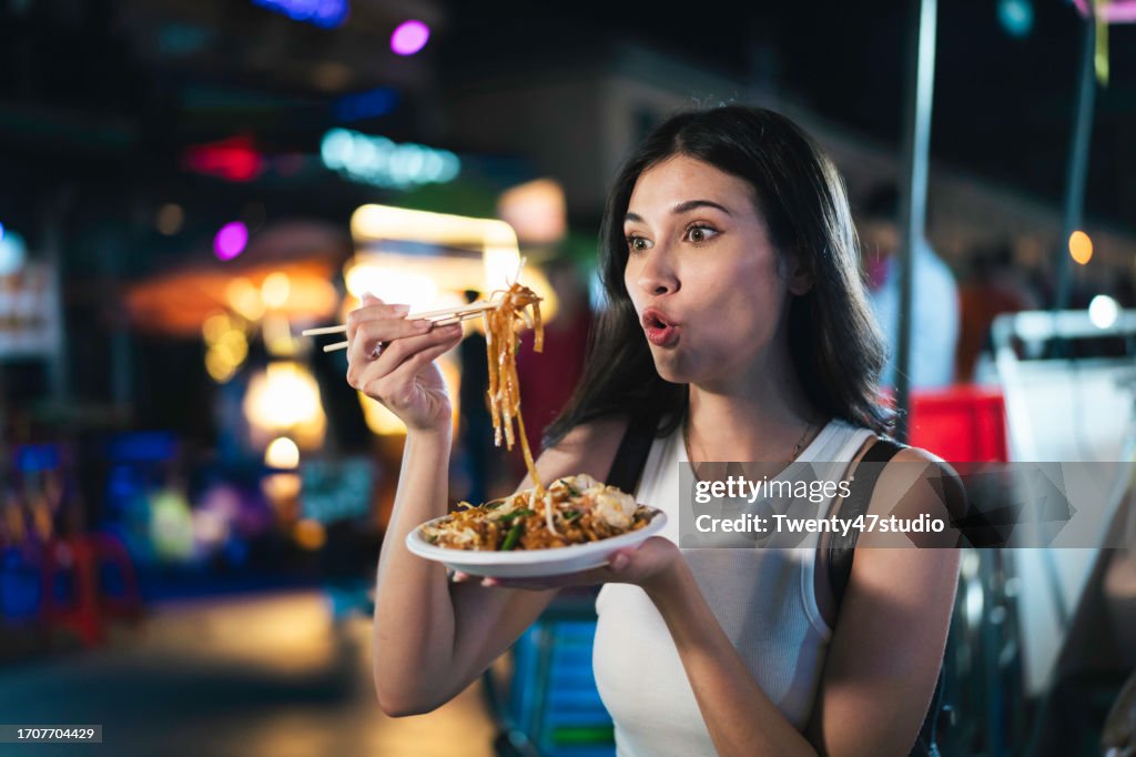 A young woman eating Pad Thai while traveling on Khao San Road at night in Bangkok, Thailand