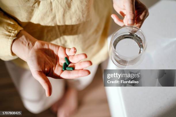 a woman standing at a table with green pills in her hand - complément vitaminé photos et images de collection