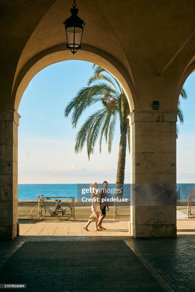Wide shot of a couple on holiday walking under an archway on the Promenade des Anglais in Nice, France