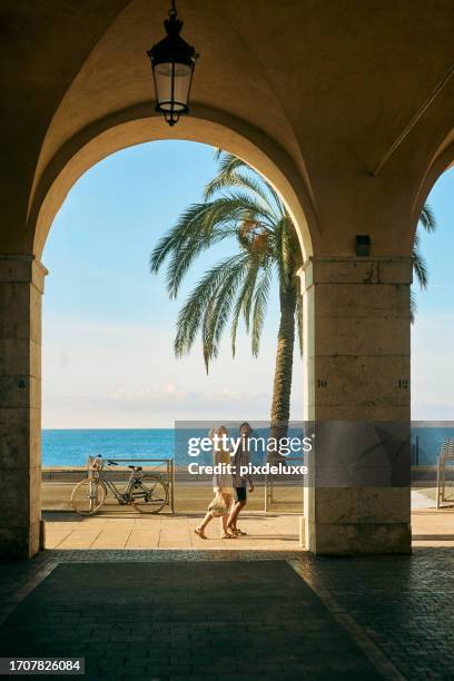 wide shot of a couple on holiday walking under an archway on the promenade des anglais in nice, france - franska rivieran bildbanksfoton och bilder