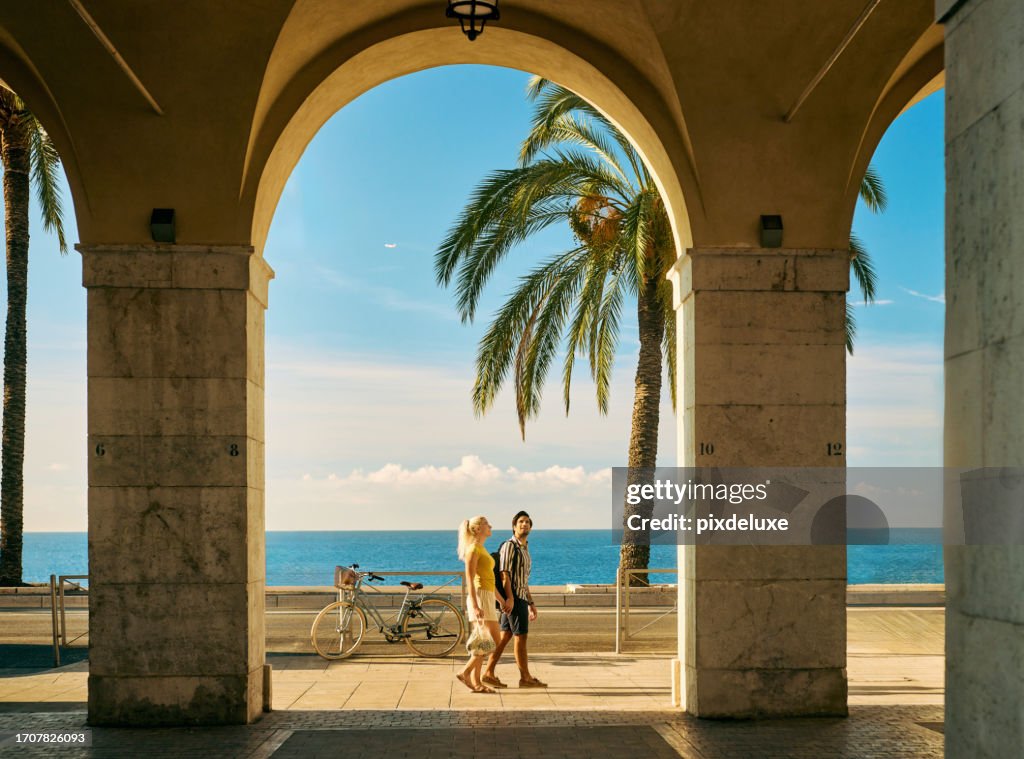 Wide shot of a couple on holiday walking under an archway on the Promenade des Anglais in Nice, France