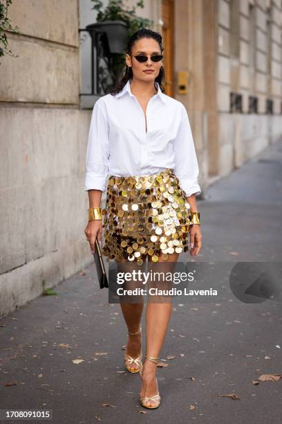 Guest wears a white shirt, gold sequins mini skirt, gold sandals, black clutch and gold bracelets, outside Rabanne, during the Womenswear...