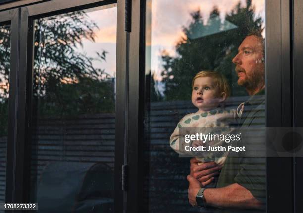 a mature father carries his baby daughter as they look through the window at the sunset - mirar-por-la-ventana fotografías e imágenes de stock