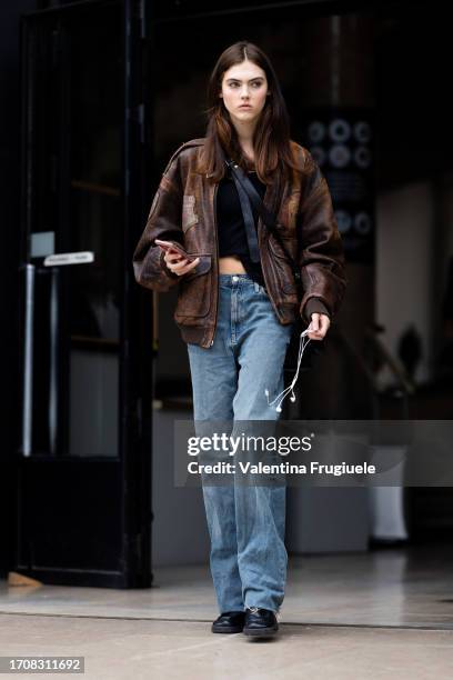 Model is seen wearing a black tank top, black leather shoes, a black cross-body bag, blue wide leg jeans and a brown leather cargo jacket outside the...