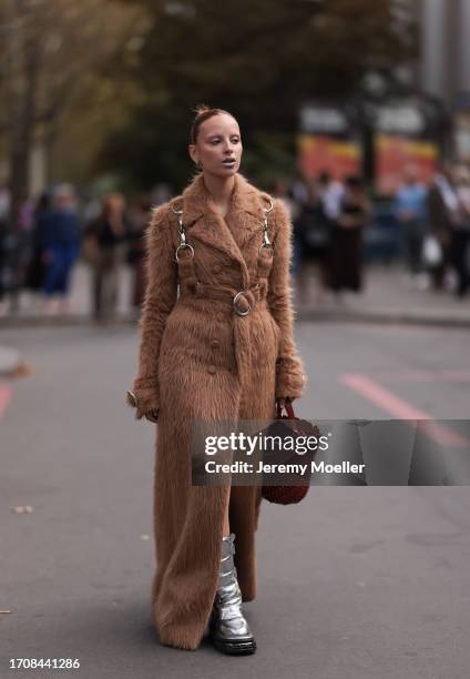 Marie Gaguech is seen wearing a long light brown coat made of fur, with silver ring details and carabiners, along with silver boots and a red leather...