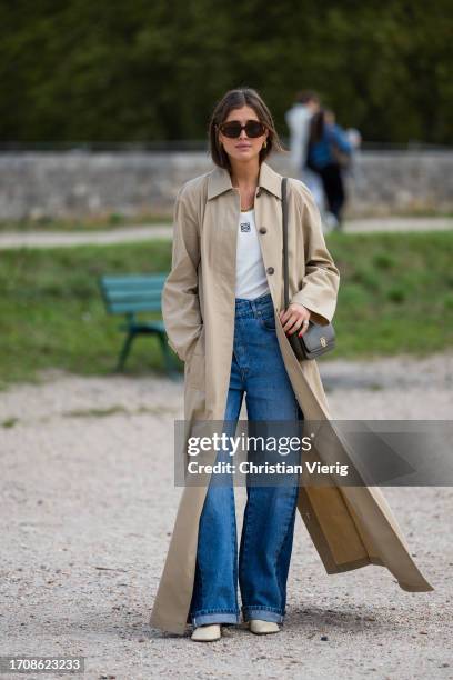 Darja Barannik wears long trench coat, denim jeans, khaki bag, white shirt, necklace, sunglasses, beige heels outside Loewe during the Womenswear...