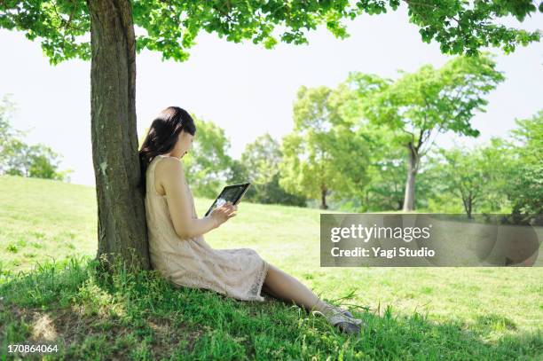 woman is using a digital tablet leaning on tree in - com sombra imagens e fotografias de stock