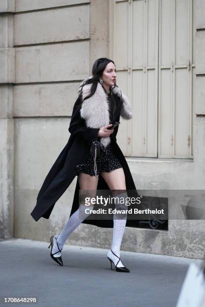 Guest wears sunglasses, earrings, a black long coat with white long fluffy collar, a balck mini skirt with printed white polka dots, white knee-high...