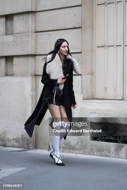 Guest wears sunglasses, earrings, a black long coat with white long fluffy collar, a balck mini skirt with printed white polka dots, white knee-high...