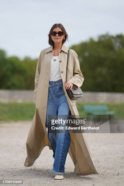 Darja Barannik wears sunglasses, a white top, a beige long trench coat, blue denim jeans / pants, outside Loewe, during the Womenswear Spring/Summer...