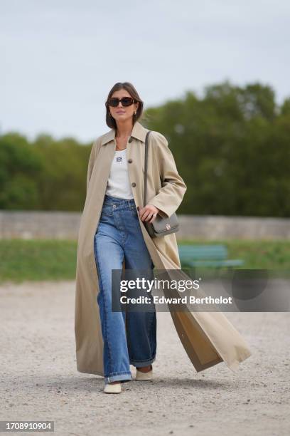 Darja Barannik wears sunglasses, a white top, a beige long trench coat, blue denim jeans / pants, outside Loewe, during the Womenswear Spring/Summer...