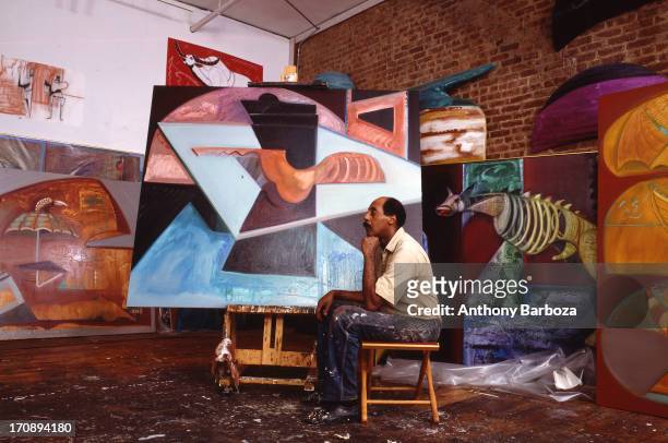 Portrait of American artist Emilio Cruz as he sits on a stool with a number of his paintings, New York, 1990s.
