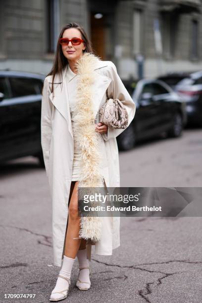 Guest wears red sunglasses, a beige top, a trench coat, a mini skirt, an ostrich feathers boa, white socks, beige studded sandals, a snakeskin bag ,...