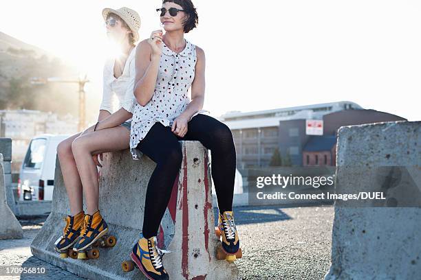 two women sitting on concrete barrier wearing rollerblades - skaten stockfoto's en -beelden