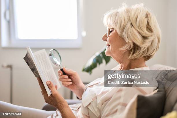 senior woman at home reading book using magnifying glass - medical dictionary stock pictures, royalty-free photos & images