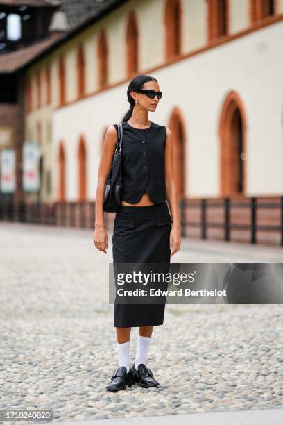 Guest wears sunglasses, earrings, a black leather bag, a black vest, a matching low waist pencil skirt, white socks, black leather moccasins with a...