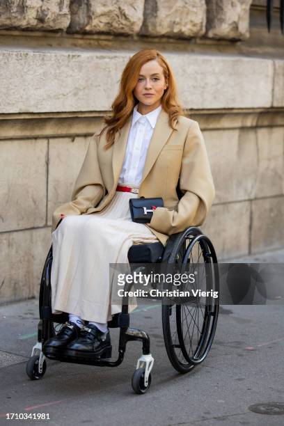 Guest in a wheelchair wears white pleated skirt, red belt, black bag, beige blazer outside Hermes during the Womenswear Spring/Summer 2024 as part of...